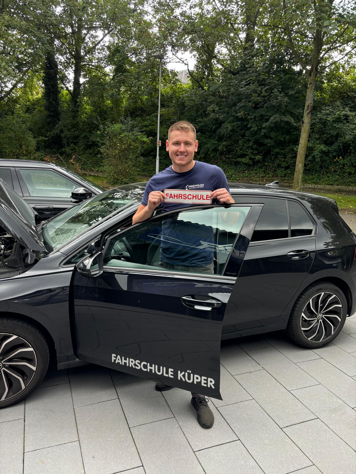 Ein lächelnder Mann mit dunkelblauem T-Shirt hält ein Fahrschulschild vor einem schwarzen Auto.